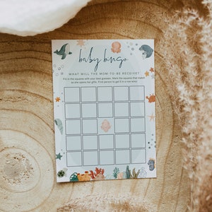 Peut inclure: Une carte de bingo pour b&eacute;b&eacute; sur le th&egrave;me de l'oc&eacute;an. La carte comporte une grille de cases avec une image d'un b&eacute;b&eacute; au centre. Le texte sur la carte indique "baby bingo" et "What will the mom-to-be receive?"
