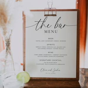 May include: A wooden bar menu sign with a copper clip, listing beers, spirits, wine, and signature cocktails. The menu features the text "the bar MENU" in elegant script. A glass of water with a lime slice is in the foreground.