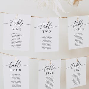 Op de afbeelding: Witte tafelkaarten met elegante kalligrafie, genummerd van één tot en met zes, opgehangen met clips aan een touwtje. Elke kaart bevat namen, ideaal voor bruiloften of evenementen. Decoratieve bloemelementen zijn zichtbaar.