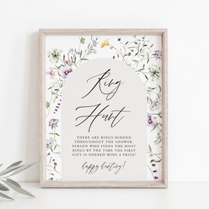 May include: A white sign with a floral border and an archway design. The text on the sign reads "Ring Hunt" and "There are rings hidden throughout the shower. Person who finds the most rings by the time the first gift is opened wins a prize! Happy hunting!"