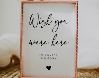 Op de afbeelding: Een roze gouden ingelijst bord met de tekst "Wish you were here" en "In loving memory" met een klein zwart hartje eronder. Het bord staat op een geweven stro mat.