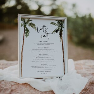 May include: A white framed menu with a watercolor illustration of palm trees. The menu lists the courses for a wedding reception: First Course, Second Course, Main Course, and Dessert. The text "Let's eat" is written in cursive at the top of the menu. The names Amalia and Hudson are listed at the bottom of the menu with the date 10-12-22.