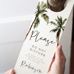 May include: White door hanger with a watercolor illustration of palm trees. The text reads "Please Do Not Disturb. We were up late celebrating the new Mr. and Mrs. Robinson."