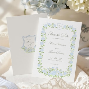 May include: Two white rectangular wedding save the date cards featuring a blue and green hydrangea floral border, a crest with the initials LT, and printed text including the names Lauren Robinson and Thomas Daniels for the date 11.28.30 in Chicago.