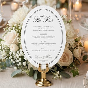 May include: A wedding bar menu card, oval-shaped with black text, is displayed on a gold stand amidst flowers. The card lists drinks, including cocktails, spirits, wine, and beer. The text reads "The Bar" and includes the names of the couple.