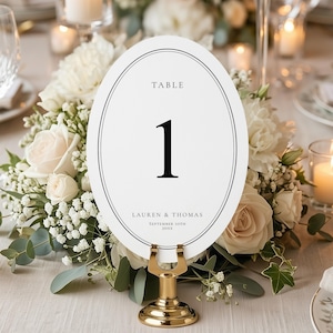 May include: An oval table number card with a black border, displaying the number 1 in a bold font. The card includes the text "TABLE" at the top and names and a date at the bottom. It is held by a gold-colored metal stand, surrounded by white flowers.