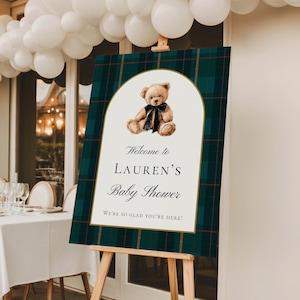 May include: A baby shower welcome sign on a wooden easel. The sign features a teddy bear illustration and text that reads "Welcome to Lauren's Baby Shower". The background has a green and navy plaid pattern.