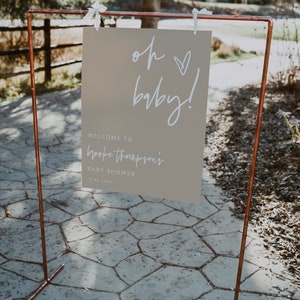 K&ouml;nnte beinhalten: Ein hellbraunes Schild mit wei&szlig;em Text, der "Oh baby!" und "Willkommen zu Brooke Thompsons Baby Shower 10-04-20XX" lautet. Das Schild h&auml;ngt an einem Gestell aus Kupferrohren.
