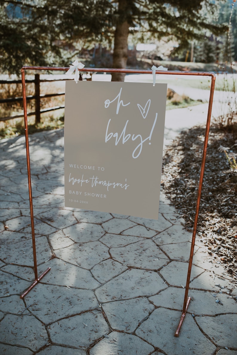 K&ouml;nnte beinhalten: Ein hellbraunes Schild mit wei&szlig;em Text, der "Oh baby!" und "Willkommen zu Brooke Thompsons Baby Shower 10-04-20XX" lautet. Das Schild h&auml;ngt an einem Gestell aus Kupferrohren.