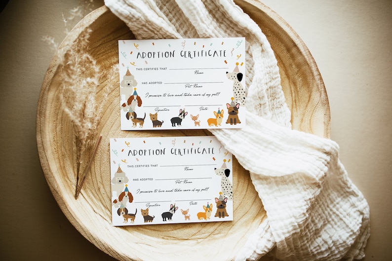 May include: Two white adoption certificates with a colorful dog theme. The certificates have spaces to fill in the name of the person adopting and the pet's name. The text on the certificate reads "Adoption Certificate" and "This certifies that... has adopted..."