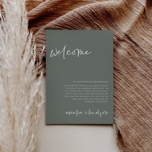 May include: A sage green welcome sign with white text that reads "Welcome to our wedding celebration, we are thrilled that you are here to share the joy of our wedding celebration. We are so fortunate to have each and every one of you in our lives and are honored that you have traveled to be with us. We hope you enjoy your stay and the weekend's festivities. Amalia + Hudson."