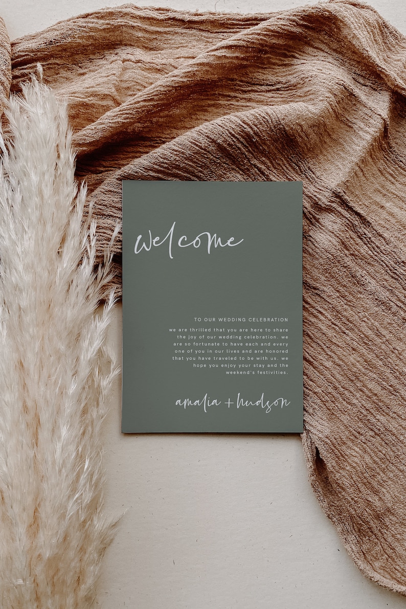 May include: A sage green welcome sign with white text that reads "Welcome to our wedding celebration, we are thrilled that you are here to share the joy of our wedding celebration. We are so fortunate to have each and every one of you in our lives and are honored that you have traveled to be with us. We hope you enjoy your stay and the weekend's festivities. Amalia + Hudson."