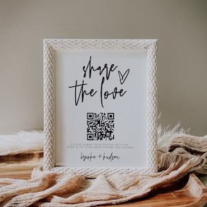 May include: A white framed sign with the words "share the love" in black script, a QR code, and the names "brooke + hudson". The sign is on a wooden tray with a beige fabric.
