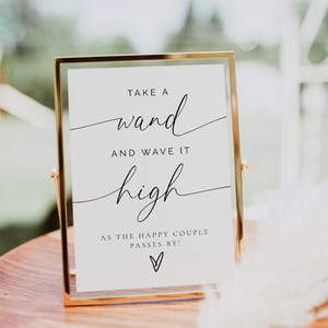 May include: A gold-framed sign with the text "TAKE A wand AND WAVE IT high AS THE HAPPY COUPLE PASSES BY!" in elegant black script. The sign is displayed on a wooden surface, with a blurred background.