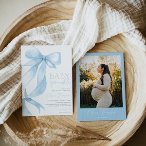 May include: Baby shower invitations with a blue bow and the words "BABY sprinkle." A photo card shows a pregnant person in a white dress. The invitations are on a wooden tray with a white cloth.