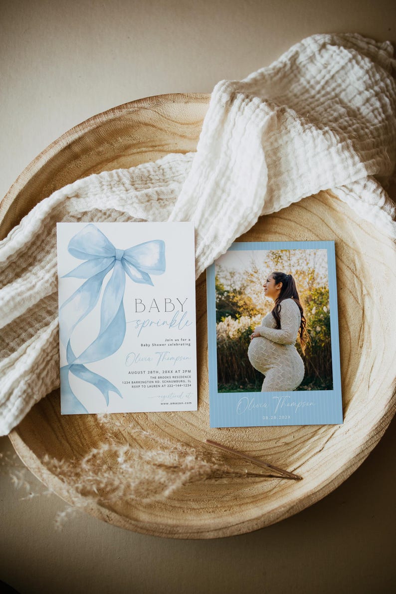 May include: Baby shower invitations with a blue bow and the words "BABY sprinkle." A photo card shows a pregnant person in a white dress. The invitations are on a wooden tray with a white cloth.