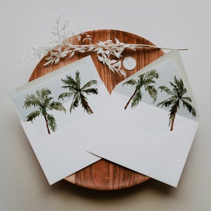 May include: Two white envelopes with watercolor palm tree illustrations are displayed on a wooden tray. The envelopes are open, revealing the palm tree design. A sprig of white dried flowers and a wax seal are also on the tray.