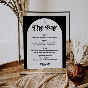 May include: A black and white sign with a gold frame that reads "The Bar" with a list of drinks available, including beer, spirits, wine, and a signature cocktail. The sign is sitting on a table with a brown tablecloth and a vase with dried flowers.