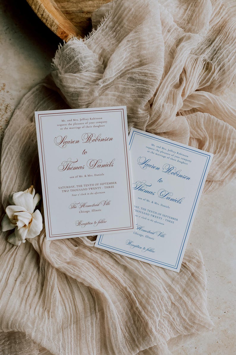 Puede incluir: Dos invitaciones de boda con caligraf&iacute;a elegante, una con borde y texto burdeos, la otra con borde azul. Las invitaciones est&aacute;n sobre una tela beige con una flor blanca. El texto dice "Lauren Robinson a Thomas Daniels".