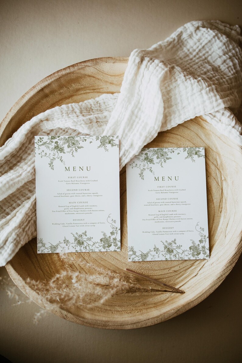 Puede incluir: Dos tarjetas de men&uacute; blancas con dise&ntilde;os florales verdes y la palabra &laquo;MEN&Uacute;&raquo; en la parte superior. Las tarjetas est&aacute;n en un cuenco de madera con un pa&ntilde;o blanco y hierba seca. El men&uacute; enumera platos como &laquo;Primer plato&raquo; y &laquo;Plato principal&raquo;.