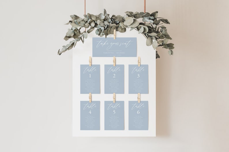 Pode incluir: Um cart&atilde;o de lugar de casamento azul claro com o texto "take your seat" e os nomes do casal. O cart&atilde;o est&aacute; pendurado em uma parede com folhagem de eucalipto e tem seis n&uacute;meros de mesa, cada um com a palavra "table" e um n&uacute;mero de 1 a 6.