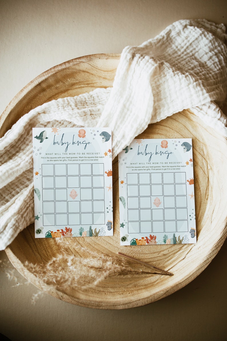 Peut inclure: Deux cartes de bingo pour b&eacute;b&eacute; bleues et blanches sur le th&egrave;me de l'oc&eacute;an. Les cartes ont une grille de cases avec des images de cr&eacute;atures marines, dont une pieuvre, une baleine, un hippocampe et une &eacute;toile de mer. Le texte sur les cartes indique "Baby Bingo" et "Que recevra la future maman ?"