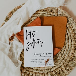 May include: A white card with black calligraphy that reads "Let's gather" and an invitation to a Thanksgiving dinner hosted by Brook & Tommy on Thursday, 26th November at 6 PM. The card is on a woven straw placemat with a brown leather envelope.