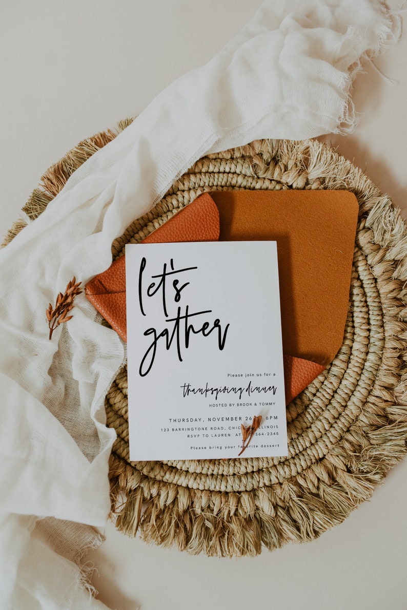 May include: A white card with black calligraphy that reads "Let's gather" and an invitation to a Thanksgiving dinner hosted by Brook & Tommy on Thursday, 26th November at 6 PM. The card is on a woven straw placemat with a brown leather envelope.