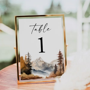 May include: A gold-framed table number sign with a watercolor illustration of a mountain range and trees. The sign reads "Table 1".