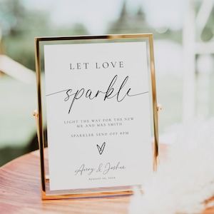 Puede incluir: Un letrero con marco dorado con las palabras "Let Love Sparkle" en una elegante escritura. El letrero incluye detalles para una boda, como los nombres "Avery & Joshua" y la fecha, 10 de agosto de 2023. El letrero se muestra sobre una superficie de madera.