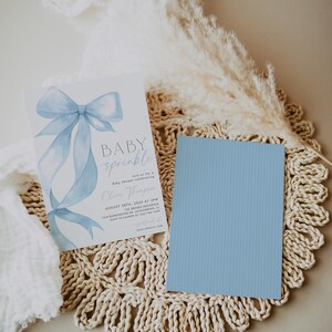 May include: A baby shower invitation with a light blue bow illustration and the words "BABY sprinkle". A light blue card is next to the invitation. The invitation includes the date, time, and location.
