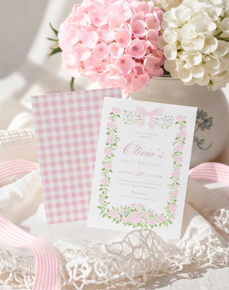 K&ouml;nnte beinhalten: Eine wei&szlig;e Einladungskarte mit rosa Blumenbord&uuml;re und Text f&uuml;r einen ersten Geburtstag. Daneben liegt eine passende rosa Vichy-Karte. Rosa und wei&szlig;e Hortensien sind im Hintergrund, zusammen mit rosa Band.