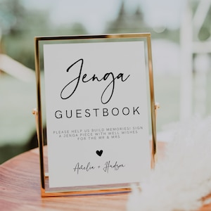 Puede incluir: Un cartel con marco dorado, fondo blanco y texto negro que dice "Jenga Guestbook". El cartel también incluye el texto "Please help us build memories! Sign a Jenga piece with well wishes for the Mr & Mrs Amelia + Hudson".