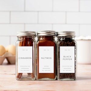 Puede incluir: Tres tarros de especias de vidrio con tapas plateadas y etiquetas blancas. Las etiquetas tienen texto negro que dice "NO. 09 CINNAMON SEASONING ALL NATURAL", "NO. 10 NUTMEG HERB ALL NATURAL" y "NO. 04 BLACK PEPPER SEASONING ALL NATURAL".