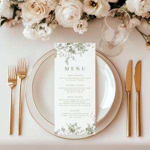 May include: A wedding menu card with a floral design in green and white, placed on a white plate with gold cutlery. The menu lists the courses for the meal. A wine glass and flowers are in the background.