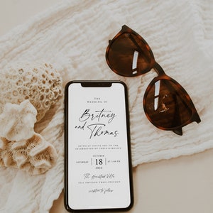May include: A wedding invitation on a smartphone screen with the text "The Wedding of Britney and Thomas" and details for the wedding ceremony and reception. The phone is lying on a white fabric surface with a pair of brown sunglasses.