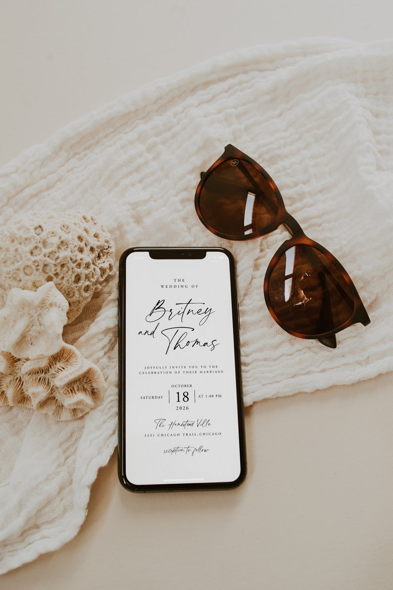 May include: A wedding invitation on a smartphone screen with the text "The Wedding of Britney and Thomas" and details for the wedding ceremony and reception. The phone is lying on a white fabric surface with a pair of brown sunglasses.