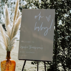 K&ouml;nnte beinhalten: Ein wei&szlig;es Schild mit dem Text "oh baby!" in kursivschrift mit einem kleinen Herz. Unter dem Text steht "Welcome to Brooke Thompson's Baby Shower 10.04.20XX". Das Schild steht auf einem Holzstaffelei in einem grasbewachsenen Bereich mit einer hohen Vase mit getrocknetem Pampasgras.