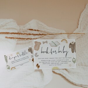 May include: Two baby shower invitation cards with illustrations of baby clothes, moons, and rainbows. One card reads "diaper raffle" and the other says "book for baby". The cards are on a white cloth background.