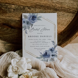 May include: A white invitation with a gold geometric frame and blue watercolor flowers. The text reads "Please join us for a Bridal Shower honoring Brooke Daniels. Saturday, December 10th, 4:00pm - 7:00pm. The Robinson's Home - 1234 Barrington Rd., Schaumburg, IL. RSVP to Lauren 222-144-1234. Hosted by Amelia, Sarah and Ericka. Registered at Amazon & Target."