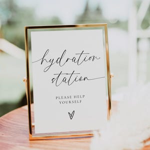 Peut inclure: Un panneau doré avec l'inscription "hydration station" en écriture élégante. En dessous, on peut lire "PLEASE HELP YOURSELF" avec un symbole de cœur. Le panneau est posé sur une surface en bois, suggérant un mariage ou un événement.