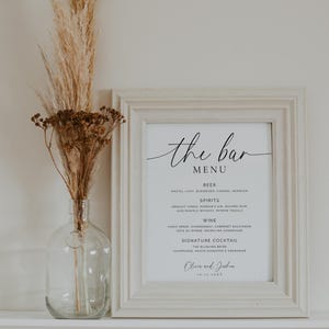 May include: A framed bar menu with a white background and black text. The menu lists beer, spirits, wine, and a signature cocktail. A clear glass vase with dried flowers sits to the left.
