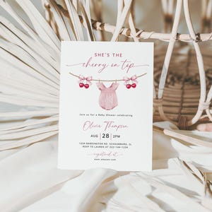 May include: A baby shower invitation with the text "She's the cherry on top" in a script font. The invitation features a pink onesie hanging on a clothesline with cherries and bows. The date and time of the shower are also included.