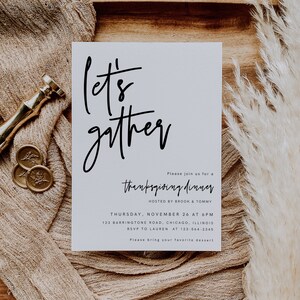 May include: A white card with black calligraphy that says "Let's gather" and an invitation to a Thanksgiving dinner hosted by Brook and Tommy. The invitation includes the date, time, address, and RSVP information. The card is on a wooden surface with a beige fabric and a dried pampas grass.