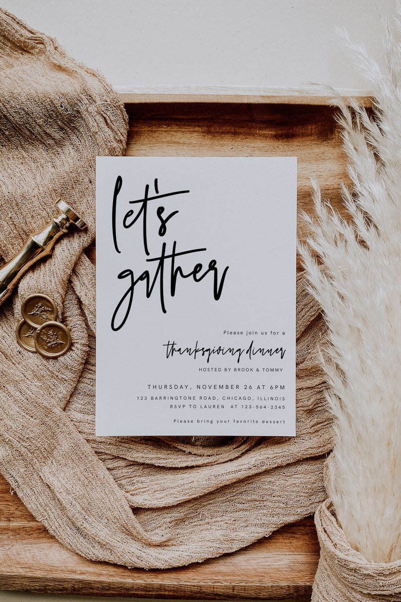 May include: A white card with black calligraphy that says "Let's gather" and an invitation to a Thanksgiving dinner hosted by Brook and Tommy. The invitation includes the date, time, address, and RSVP information. The card is on a wooden surface with a beige fabric and a dried pampas grass.