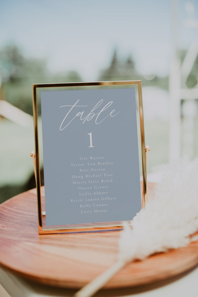 Pode incluir: Um cart&atilde;o de n&uacute;mero de mesa com moldura dourada e fundo azul claro. O n&uacute;mero "1" &eacute; exibido em branco, juntamente com uma lista de nomes em branco: Teri Watson, Tyrone Tom Bradley, Ross Harvey, Doug Michael Tran, Mattie Steve Brock, Sharon Garner, Leslie Abbott, Kristi Lauren Gilbert, Kelly Conner, Carla Munoz.