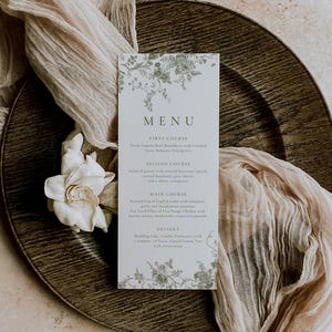 Puede incluir: Una tarjeta de men&uacute; de boda con detalles florales, que muestra la palabra "MENU" en la parte superior. La tarjeta est&aacute; colocada sobre un plato de madera con tela drapeada y una flor blanca. El men&uacute; enumera los platos.