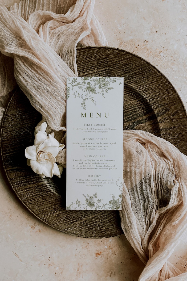 Puede incluir: Una tarjeta de men&uacute; de boda con detalles florales, que muestra la palabra "MENU" en la parte superior. La tarjeta est&aacute; colocada sobre un plato de madera con tela drapeada y una flor blanca. El men&uacute; enumera los platos.