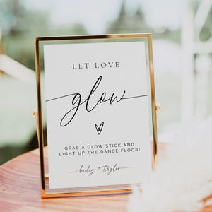 Può includere: Un cartello con cornice dorata con le parole "LET LOVE glow" in elegante scrittura. Sotto, si legge "GRAB A GLOW STICK AND LIGHT UP THE DANCE FLOOR!" con i nomi "bailey + tayler" in basso. Il cartello è su una superficie di legno.
