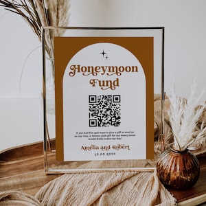 May include: A framed sign with a brown background and white text that reads "Honeymoon Fund". A QR code is displayed below the text. The sign also includes the text "If you had fun and want to give a gift to send us on our way, a Venmo cash gift for our honeymoon would really make our day! Amelia and Robert 10.06.2026".